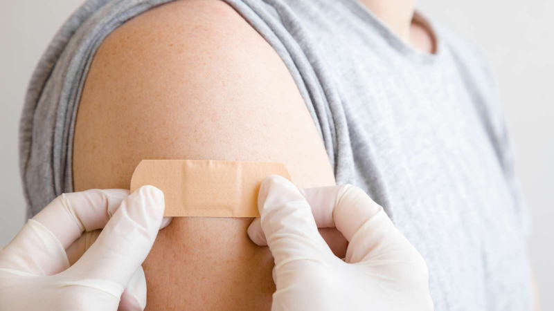 Photo of medical professional with gloves on placing a plaster on patients upper arm.