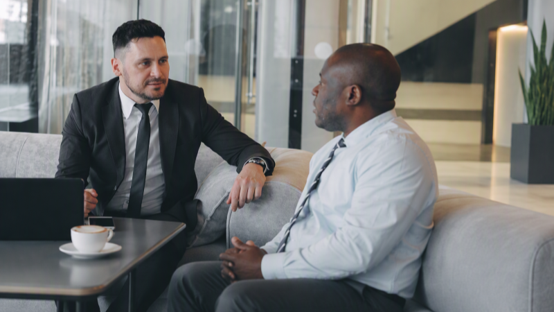 Two men in business attire talking in an office with laptop, phone and coffee on the table.
