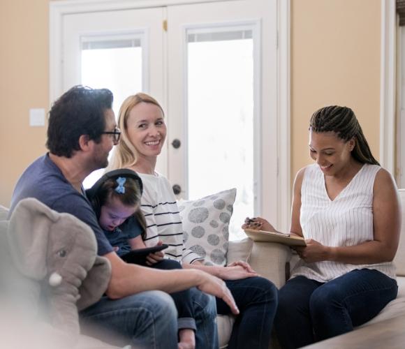 A family of three sitting together on a couch talking to a social worker sitting across from them and making a note on a clipboard.