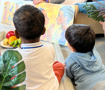 Two children sitting on the carpet looking at book being presented by an adult.