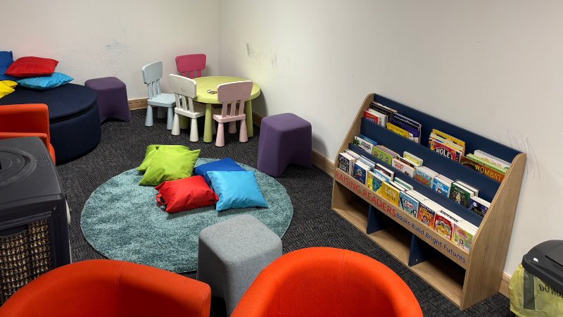 image of the interior of the book nook room, showing a fluffy blue floor rug with pillows stacked on top in various colours, two large red reading chairs, an A/C unit, large navy blue ottoman with pillows on top, a pastel coloured child's table and chairs