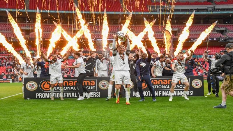 Photo of Bromley FC players celebrating with the trophy on the pitch at Wembley, with fireworks in the background.