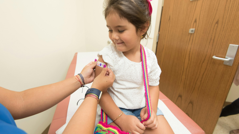 Photo of a young girl having a plaster stuck to their arm by a medical professional while sat down.