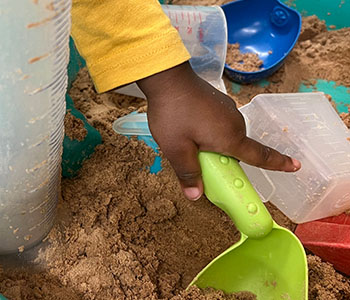 Child’s hand holding green spade, scooping sand in sand box.