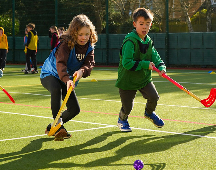 Munchkin Sports - Multiple Schools: Photo of two children playing hockey together outside on a pitch.