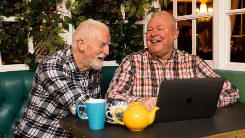Two older men sat talking together at a table over a pot of tea.