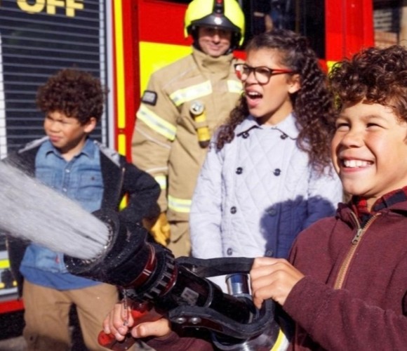 Young person holding a fire hose with firefighter, fire truck and other residents in the background.