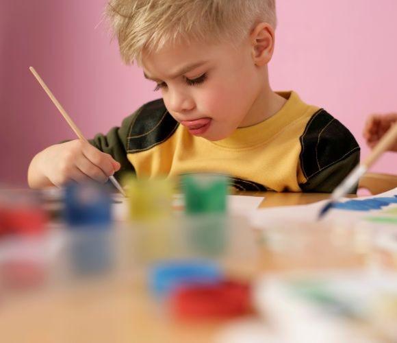 Image shows a young boy painting using different coloured pots of paint.