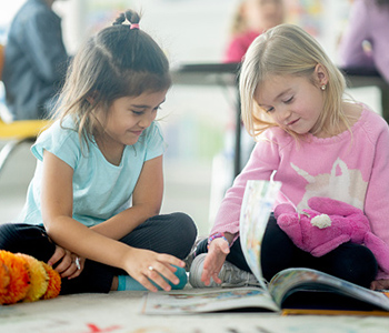Two young children sitting together indoors, looking at a book.