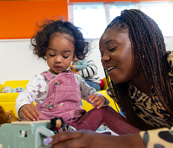 Child playing with toys alongside a childcare worker in a day nursery.